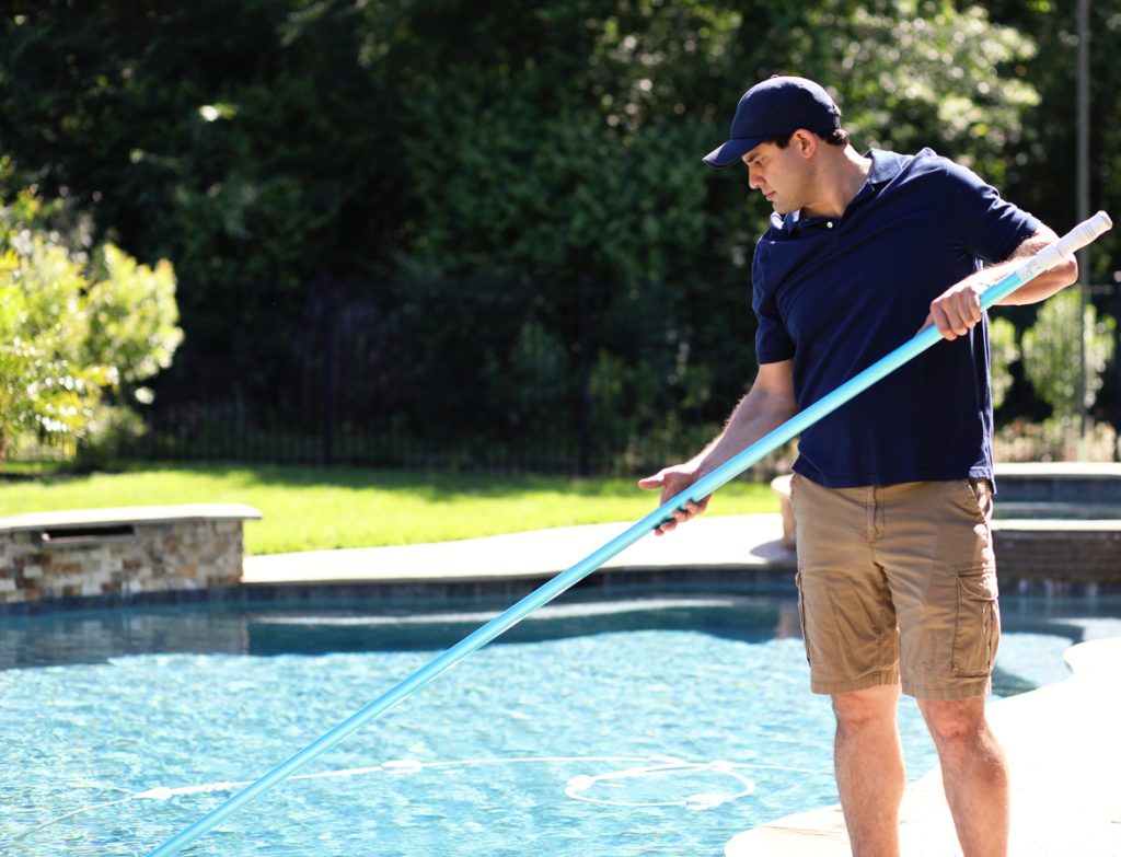 Man in a navy shirt and cap cleaning an inground pool with a skimmer pole on a sunny day, surrounded by greenery and a stone deck.